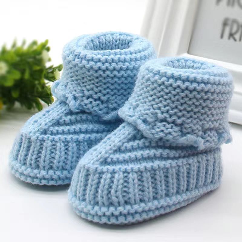 Pair of blue knitted baby booties on a white surface with a plant in the background.