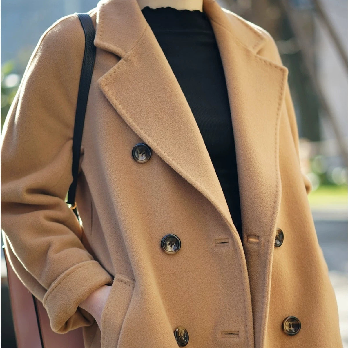 A close-up of a person wearing a camel-colored double-breasted coat over a black top, with a black shoulder bag strap visible against a softly blurred outdoor background.