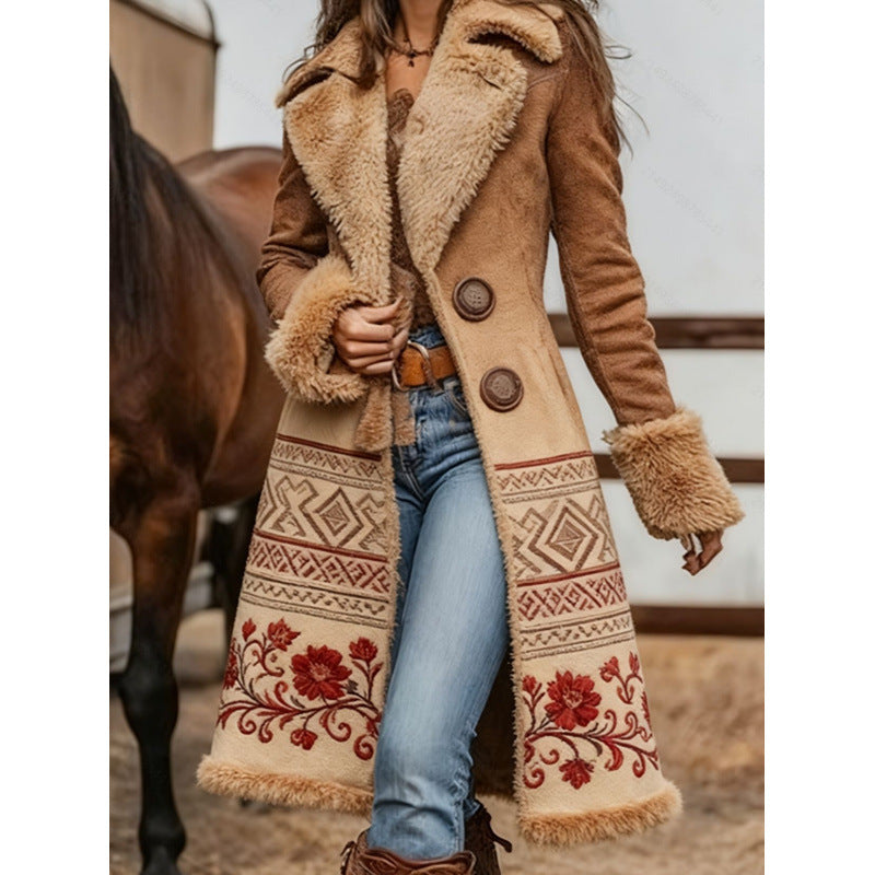 A woman wearing a long brown suede-like coat with a beige faux-fur collar and cuffs, double-breasted buttons, and a cream patterned lower panel with red floral embroidery, paired with blue jeans in an outdoor barn setting with a horse visible.