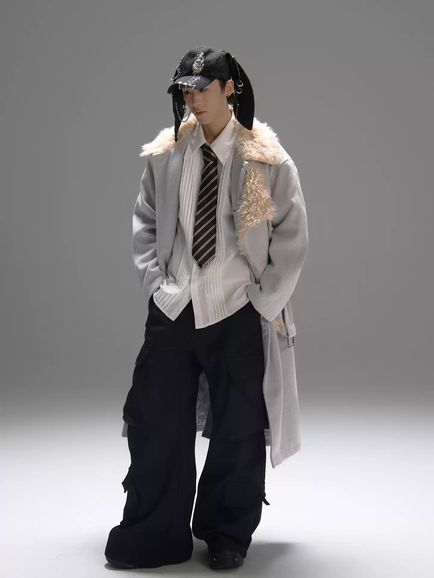 Person wearing a long coat with fur collar, tie, and black pants on a gray background
