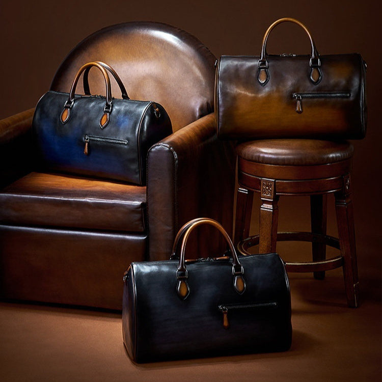 Three leather duffel bags in dark blue, brown, and black are arranged on and around a brown leather armchair and matching stool against a warm brown studio background.