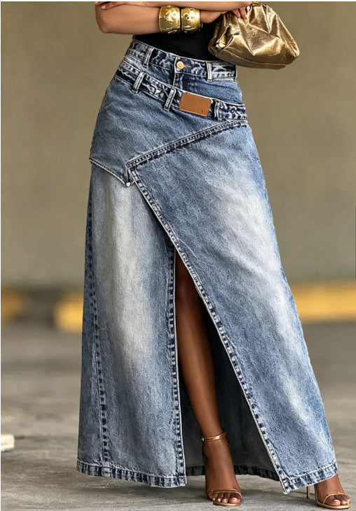 A woman wearing a long light-wash denim maxi skirt with a diagonal front slit, styled with a black top, gold bracelets, a metallic gold clutch, and brown heeled sandals.