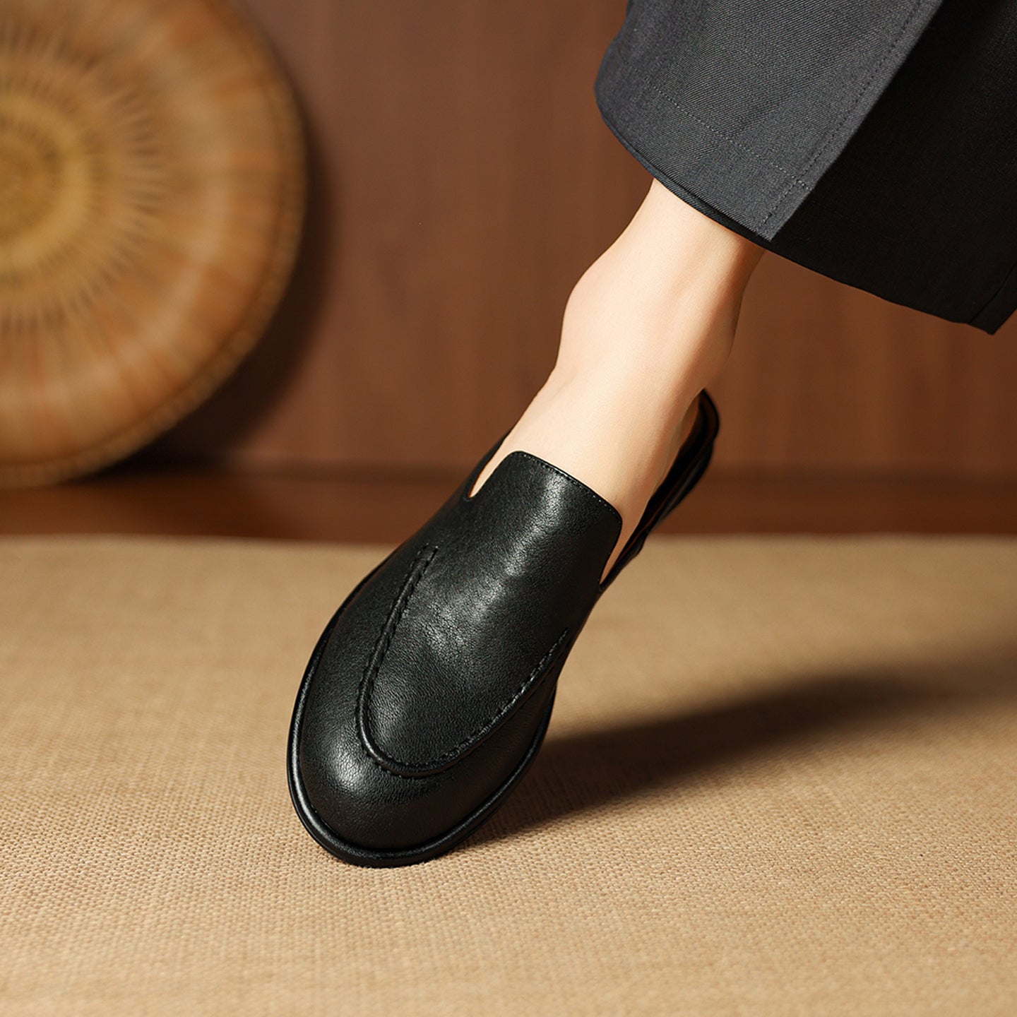 Black leather slip-on shoe worn by a person on a beige surface with a woven mat in the background.