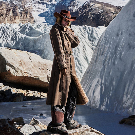 Person in a long coat and hat standing in a snowy landscape with mountains.