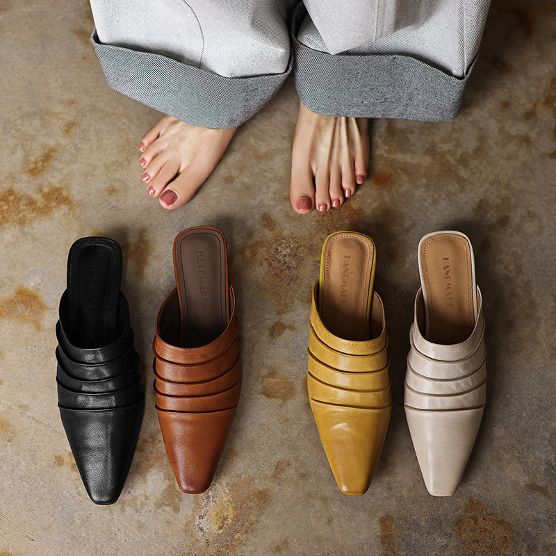 Four pairs of pointed-toe shoes in black, brown, yellow, and beige on a textured floor.