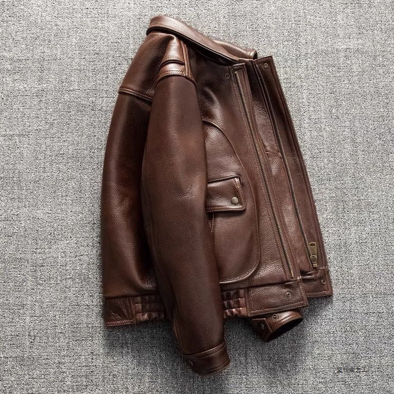 A brown leather jacket with a front zipper, flap pocket, and ribbed cuffs and hem, laid flat on a textured gray background.