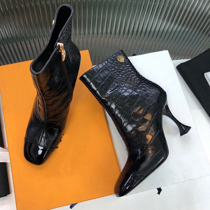 Black crocodile-patterned high-heeled boots on a white surface with an orange box.