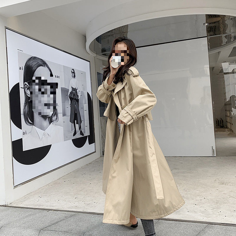 Person wearing a beige trench coat in an indoor setting with a large mirror and artwork on the wall. by millivan