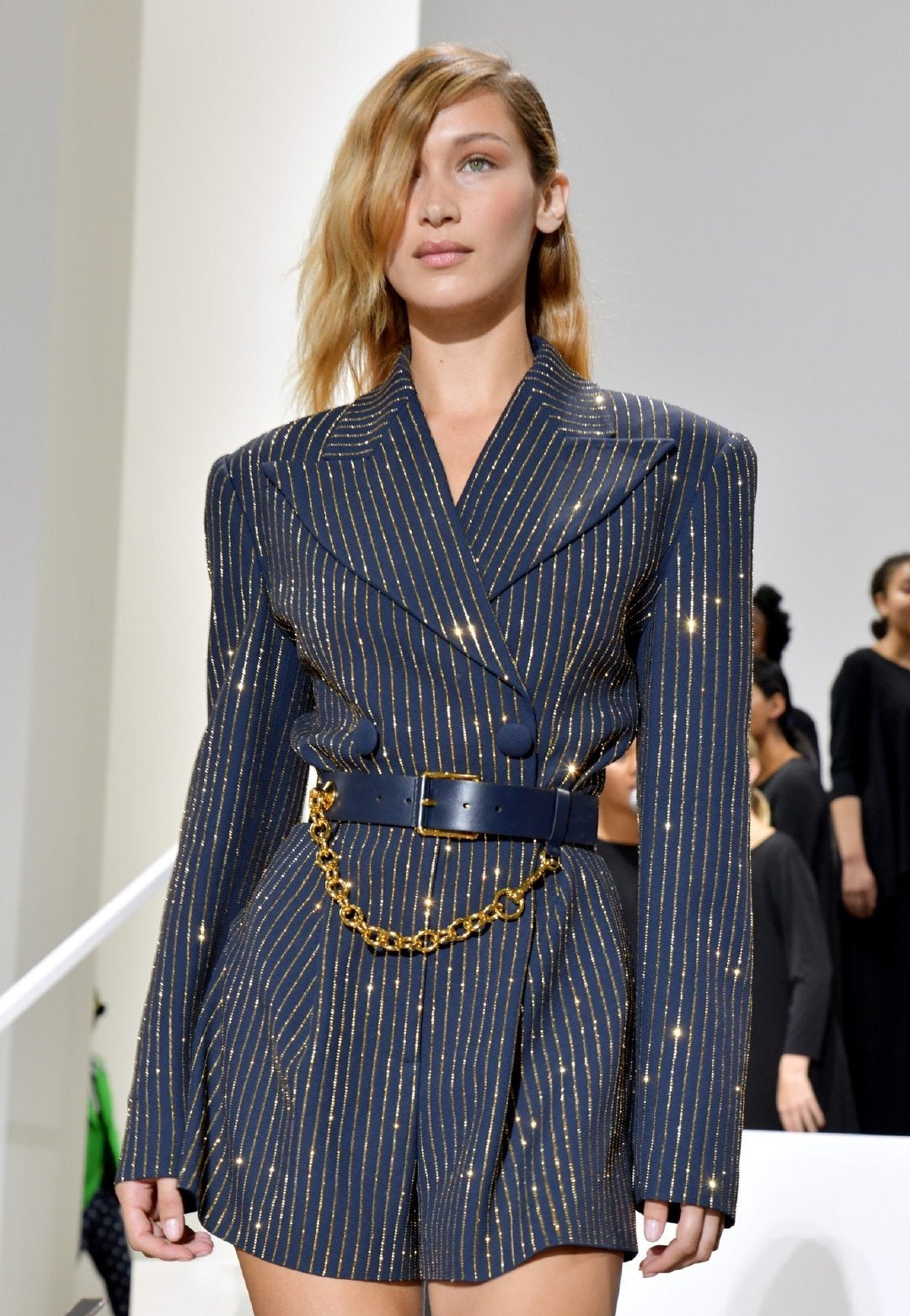 Model wearing a blue pinstripe suit with a gold chain belt on a runway. millivan