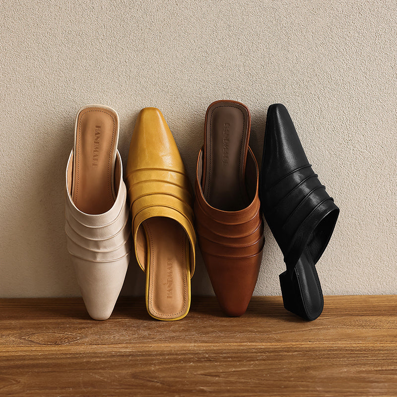 Four pairs of mules in white, yellow, brown, and black on a wooden surface with a beige wall background.