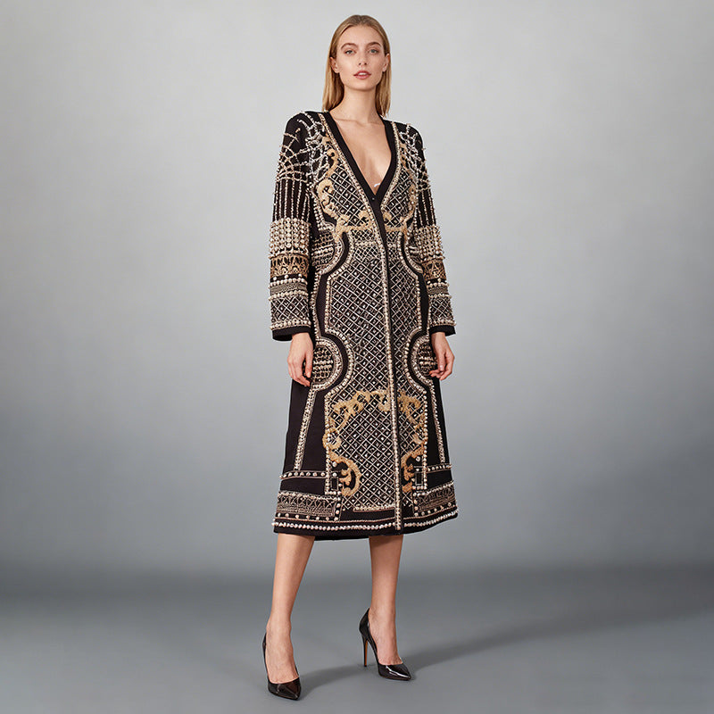 A model wears a long-sleeved, knee-length black cardigan featuring intricate gold and beige geometric and scroll patterns, paired with black heels against a neutral gray studio background.