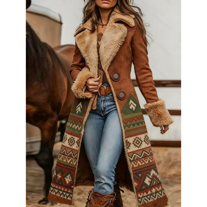 A woman models a long brown suede-like coat with a beige shearling collar and cuffs, featuring a colorful geometric patterned panel on the front and back. She is wearing blue jeans and brown boots, standing in front of a horse in a rustic indoor setting.