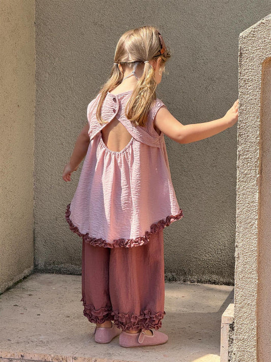 Child wearing a pink outfit with ruffled details, standing against a plain wall.