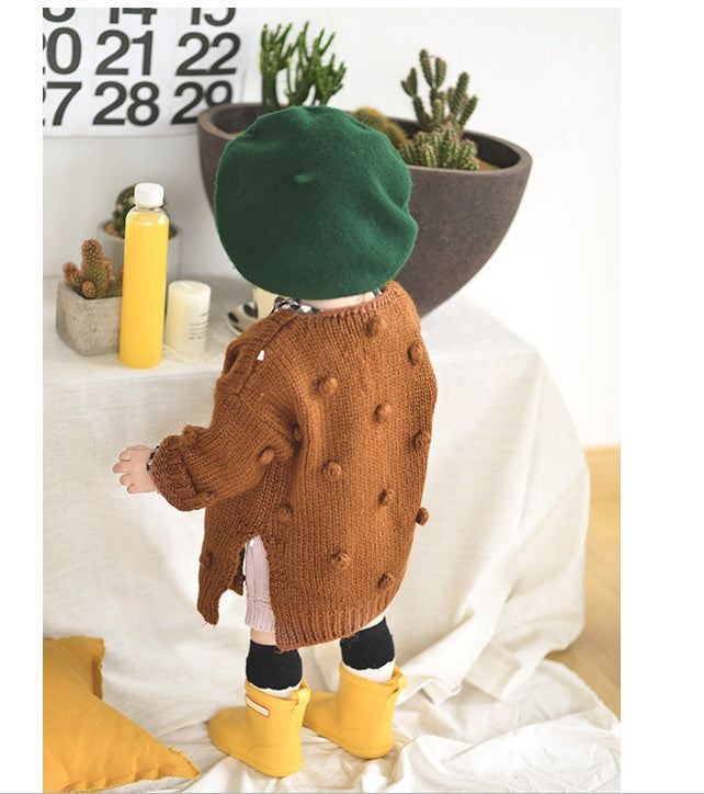 Child wearing a brown coat with button details, green hat, and yellow boots in a room setting.