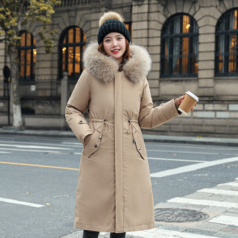 Woman in a long beige coat with a fur-lined hood standing on a street holding a coffee cup.