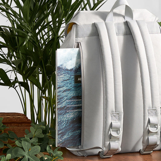Gray backpack with a side pocket displaying an ocean view, placed among plants.