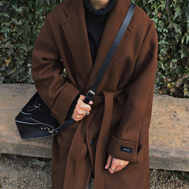 Person wearing a brown coat holding a black bag against a green leafy background