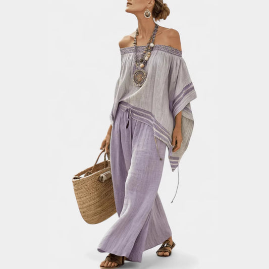 Woman wearing a light gray off-shoulder top and purple pants, holding a straw bag on a white background.