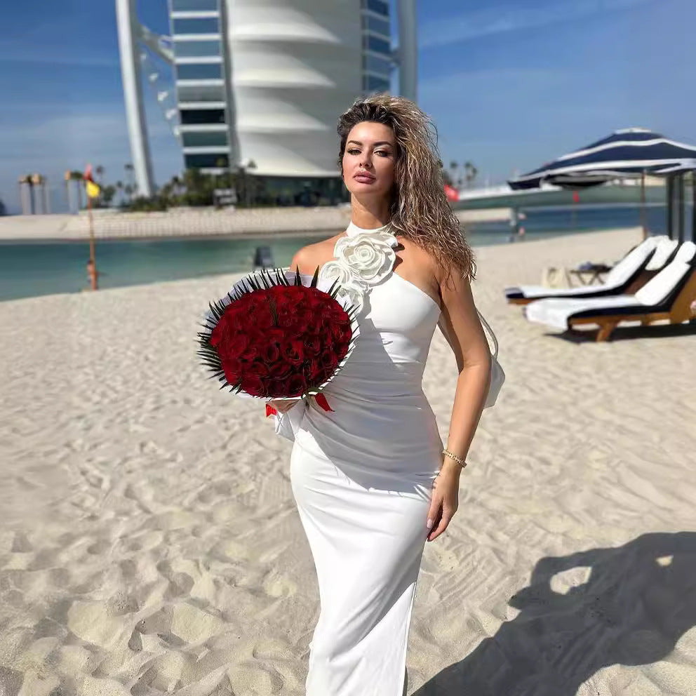 Woman in a white dress holding a red flower bouquet on a beach with a building in the background
