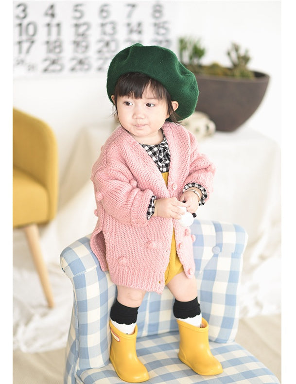 Child wearing a pink coat, green beret, and yellow boots in a cozy indoor setting.