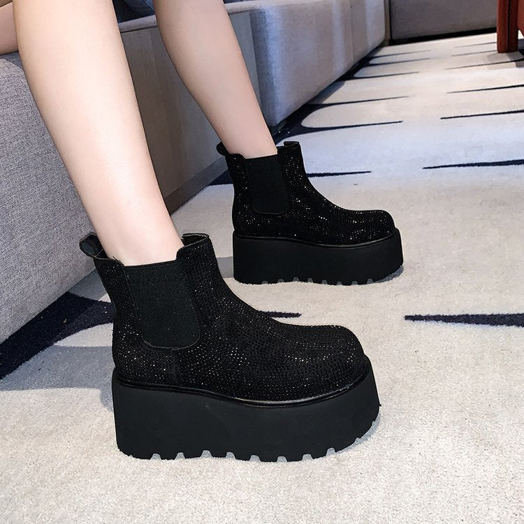 Black platform Chelsea boots covered in sparkling rhinestones, shown on a model's feet in a modern indoor setting with a gray sofa and patterned carpet.