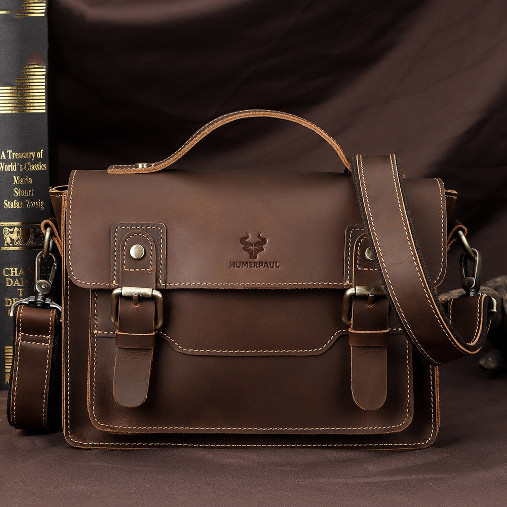 A brown leather messenger bag with a top handle, front flap, buckle details, and a detachable shoulder strap, displayed against a dark brown fabric backdrop with a book partially visible on the left.