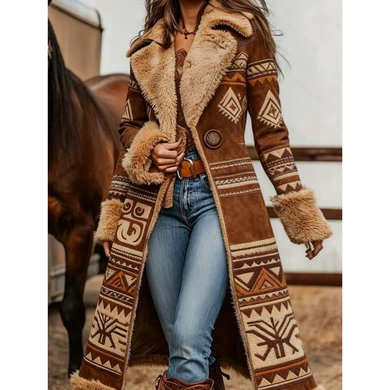 A woman wearing a long brown suede-like coat with cream and tan Southwestern geometric patterns and fluffy faux-fur trim, paired with blue jeans and brown boots, standing in front of a horse and stable background.