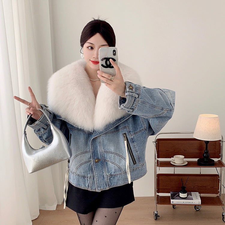 Woman wearing a denim jacket with a fur collar, holding a phone and a silver handbag indoors.