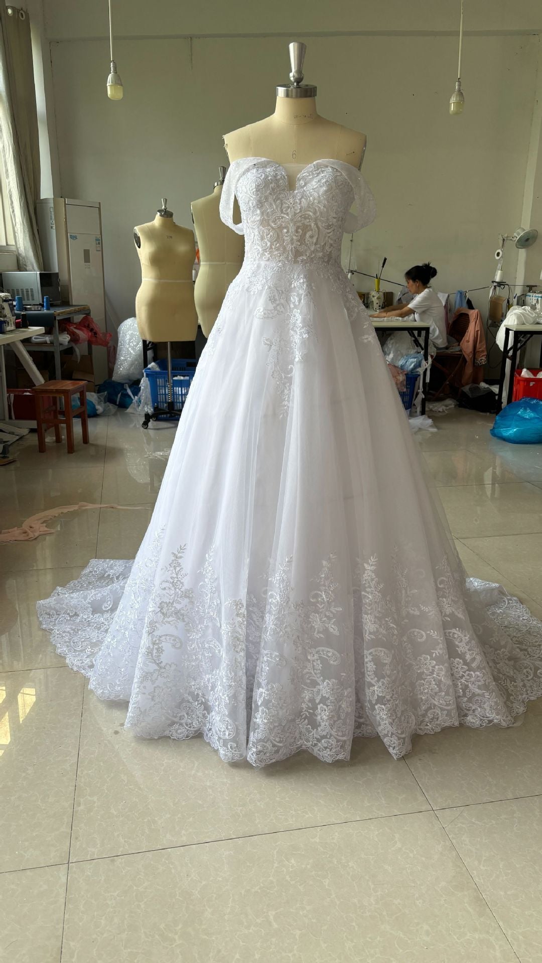 White lace wedding dress on a mannequin in a room with other mannequins and furniture.