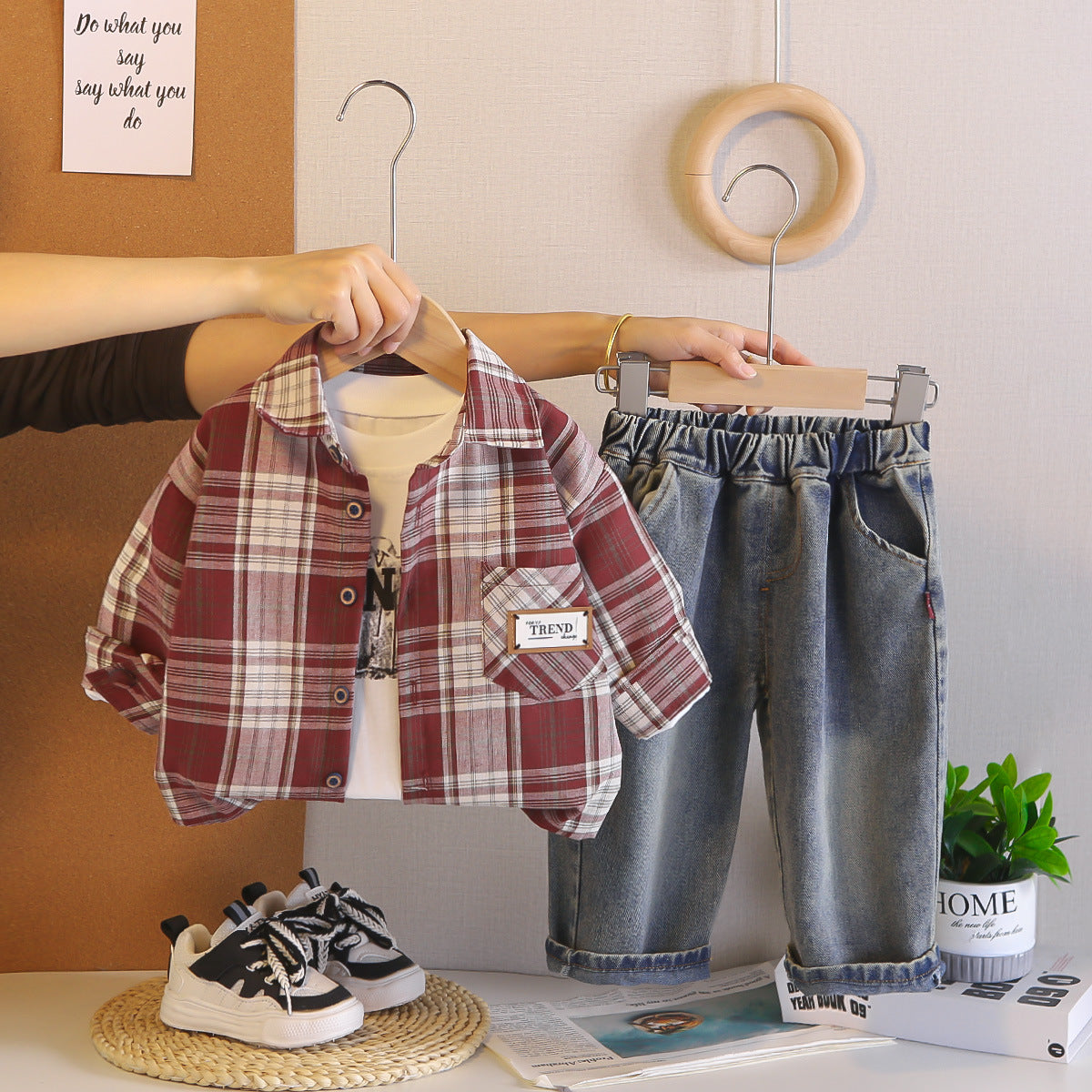 Child's plaid shirt, jeans, and sneakers on hangers with a plant and magazine in the background.