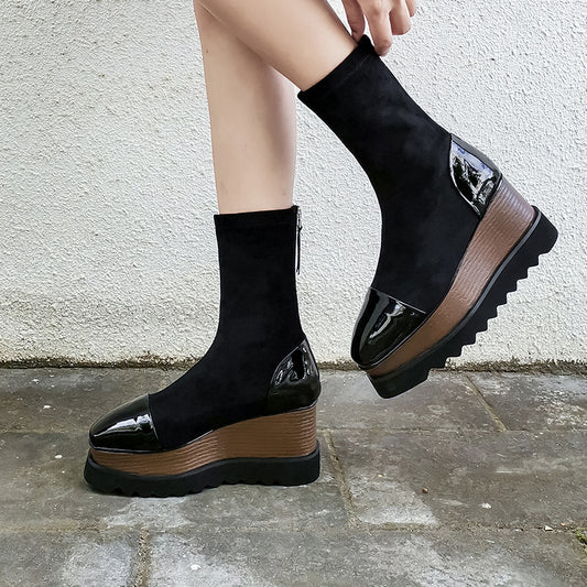 Black platform ankle boots with brown soles worn by a person against a white wall.