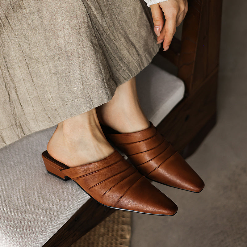 Brown leather mules worn by a person sitting on a chair.