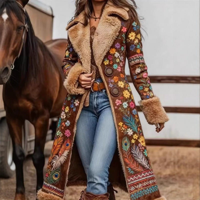 A woman wearing a long brown suede-like coat with colorful floral and feather embroidery, faux-fur trim, and large buttons, standing outdoors next to a horse.