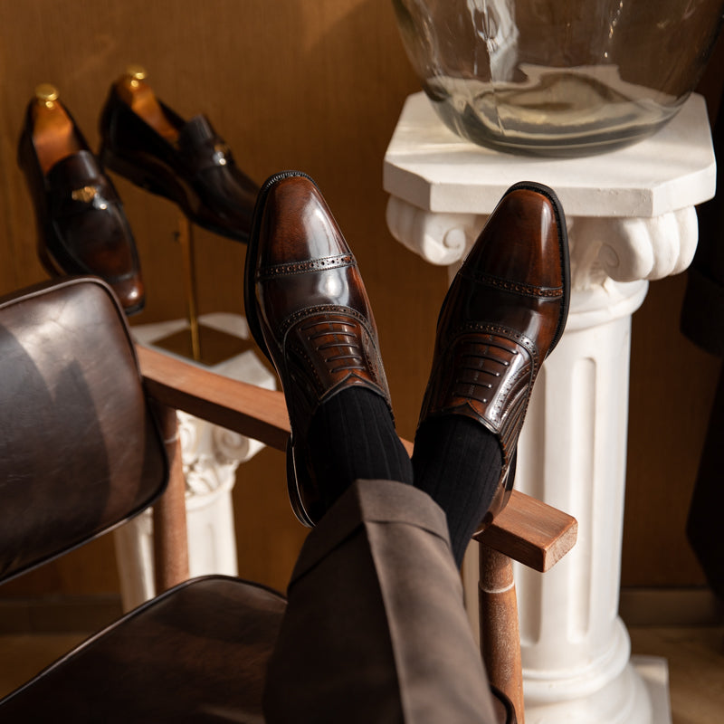 Person wearing brown dress shoes and black socks sitting on a chair with a decorative background.