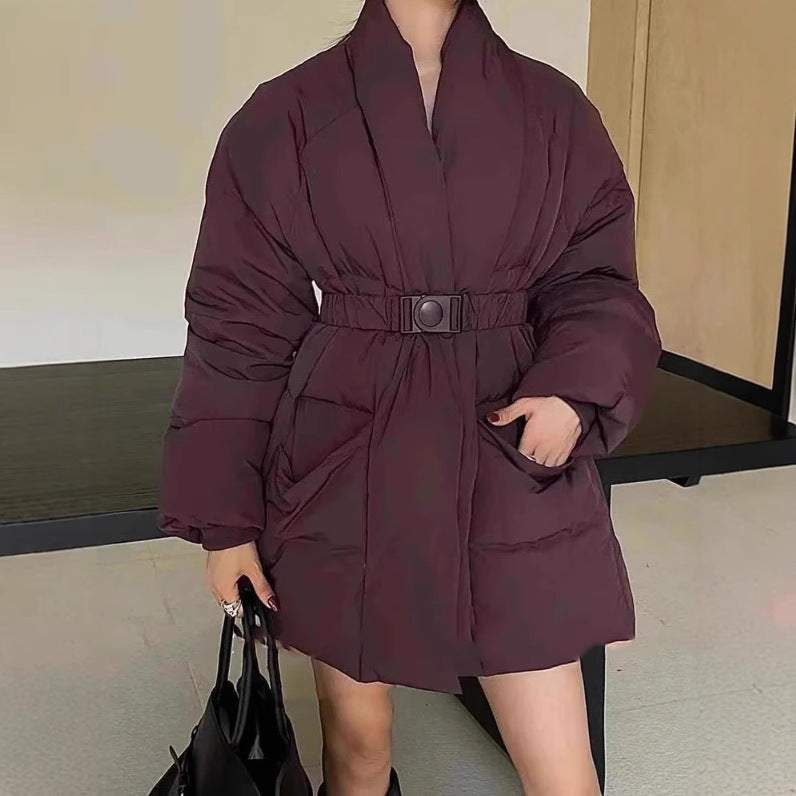 Person wearing a maroon puffer coat with a belt, holding a black bag indoors.