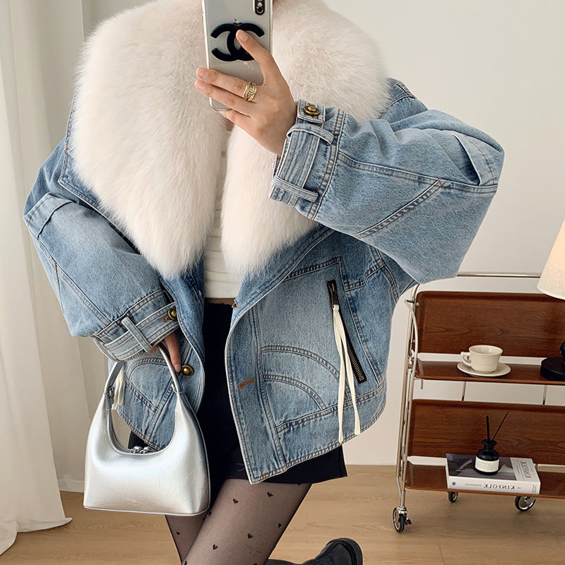 Person wearing a denim jacket with a white fur vest, holding a phone, in a room with a side table and lamp.