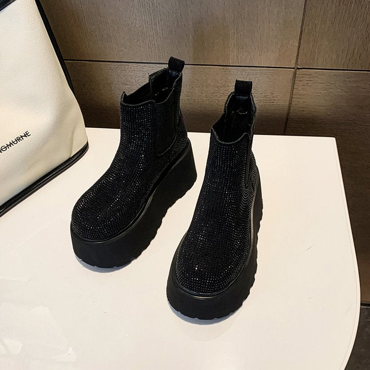 A pair of black platform ankle boots covered in sparkling rhinestones, displayed on a white table next to a white handbag.