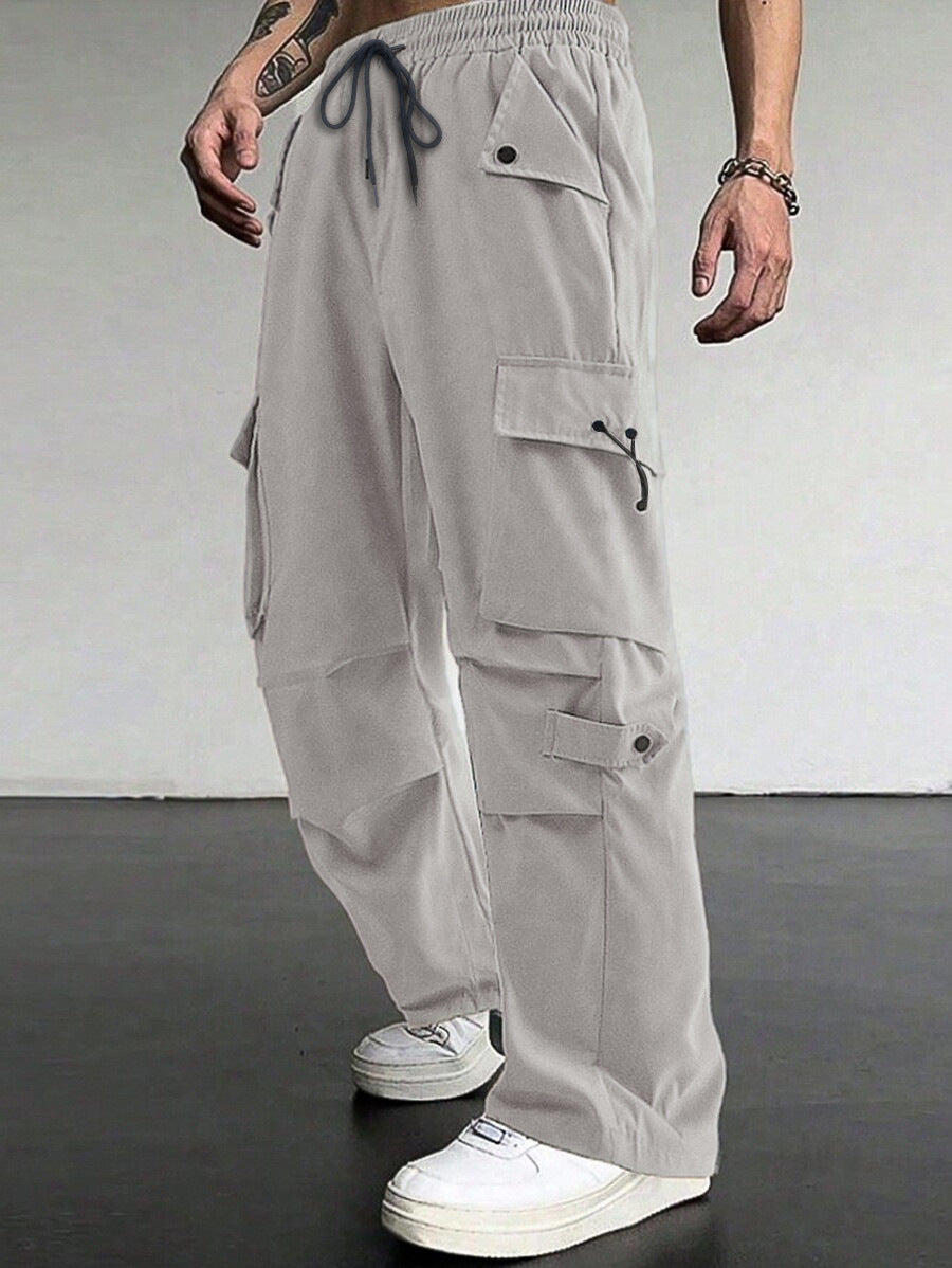 A person wearing light gray cargo pants with multiple flap pockets, an elastic drawstring waist, and wide legs, paired with white sneakers against a plain indoor wall and dark floor.