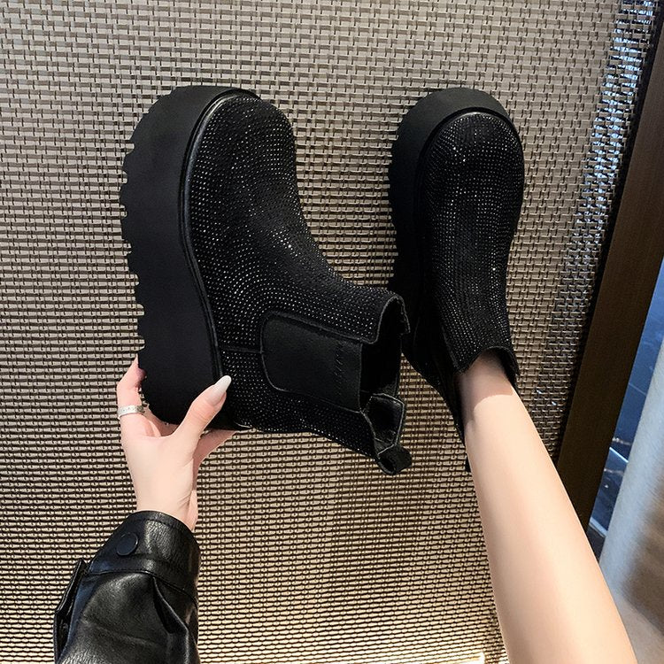 Black rhinestone-embellished ankle boots with chunky platform soles, shown with one boot being held by a hand and the other worn on a foot, set against a woven brown textured floor.