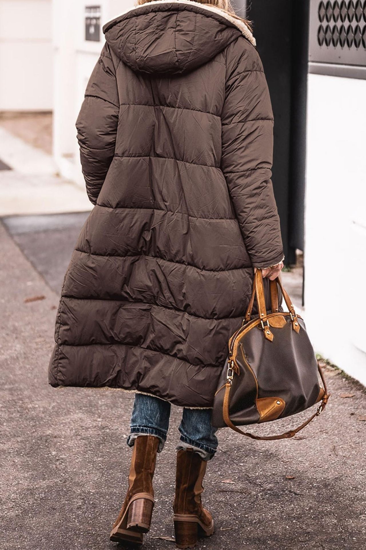 Person wearing a long dark brown puffer coat and holding a brown leather bag on a sidewalk.