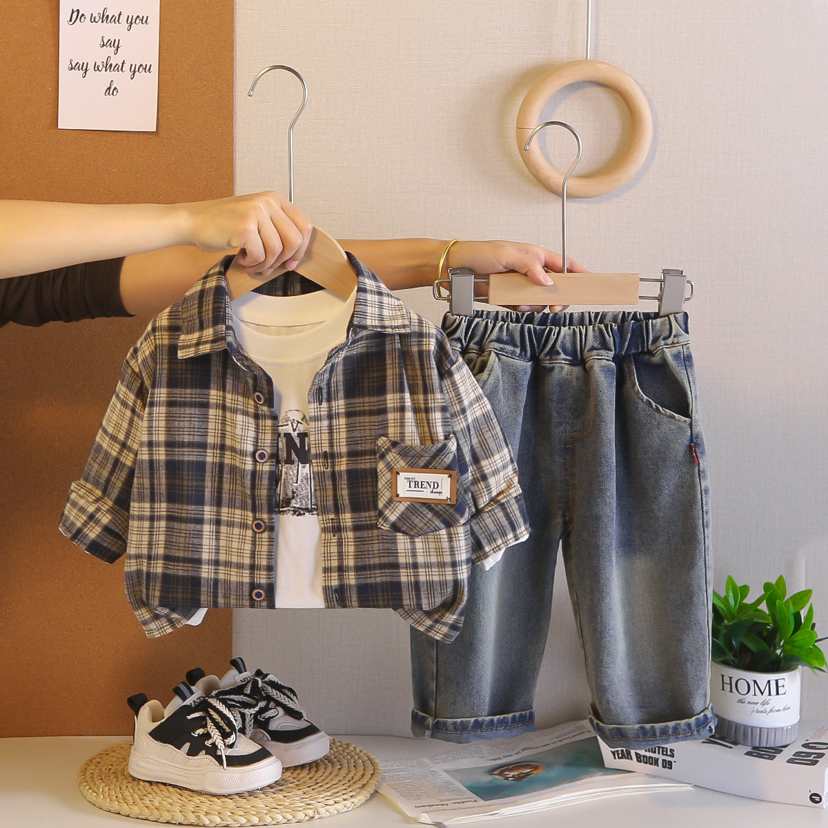 Children's plaid shirt, jeans, and sneakers on hangers with a plant and magazine in the background.