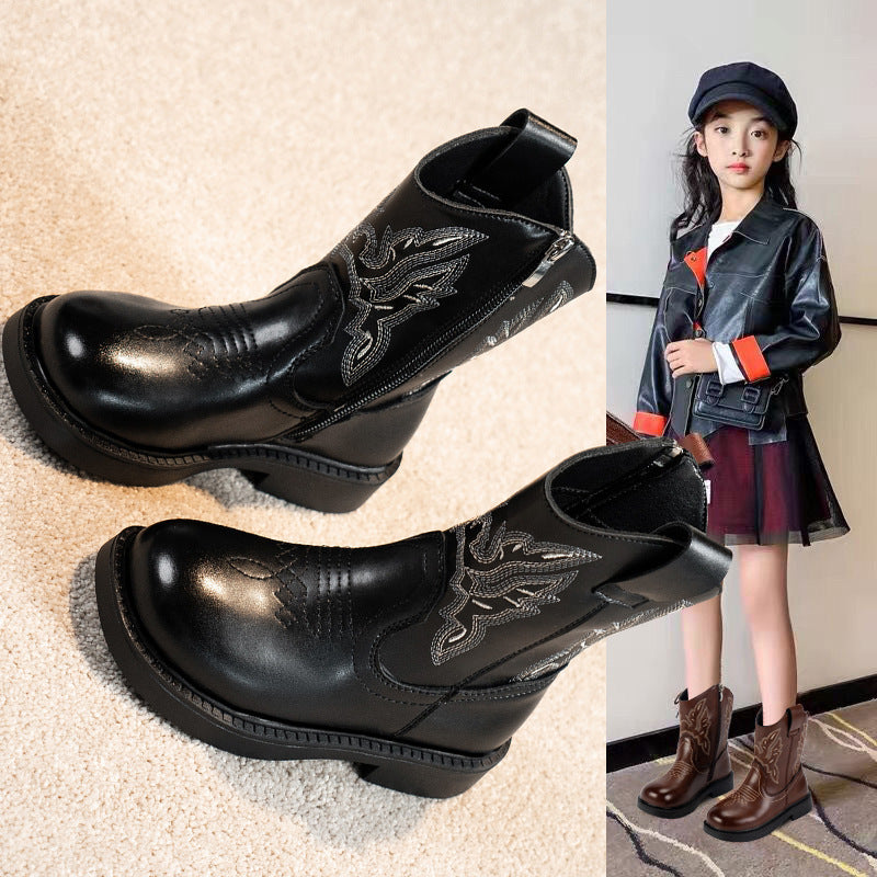 Black leather boots with embroidery design on a beige surface, next to a person wearing a jacket and skirt.