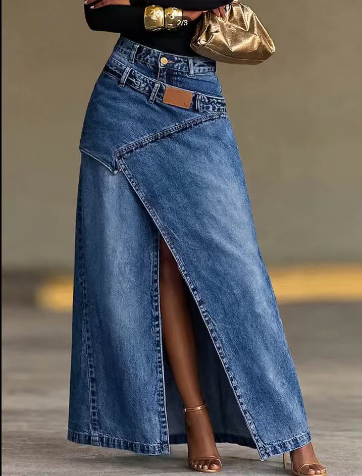 A high-waisted blue denim maxi skirt with an asymmetrical wrap-style front and a thigh-high slit, shown on a model holding a metallic gold clutch.