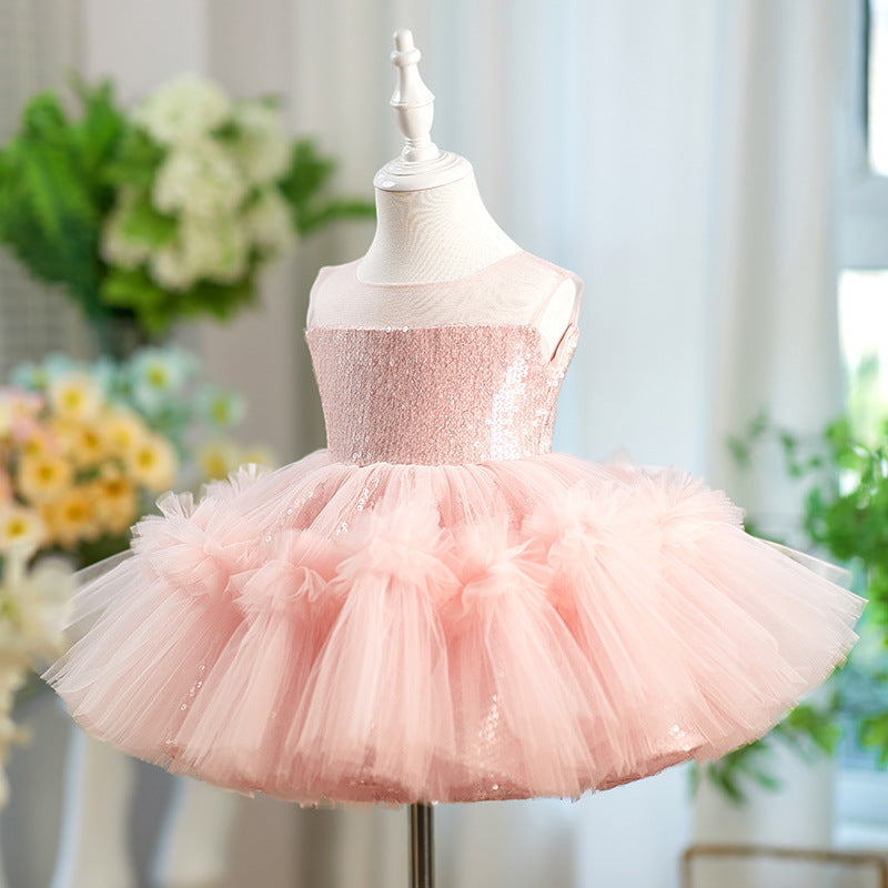 A pink sequined tutu dress displayed on a white mannequin dress form, set against a softly blurred indoor background with flowers and greenery.