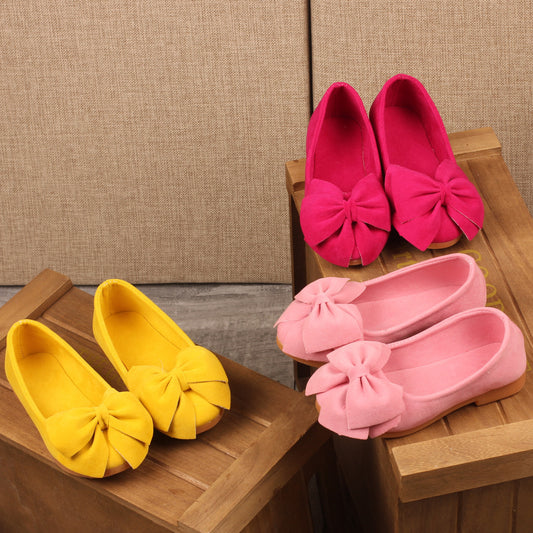 Yellow and pink shoes with bow details on wooden stools against a beige textured wall.