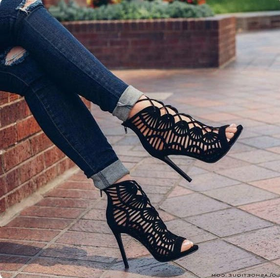 Black laser-cut high-heeled shoes worn with ripped jeans on a paved surface.