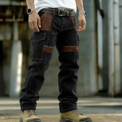 A person wearing dark corduroy cargo pants with brown leather patch pockets and brown contrast panels, paired with a white shirt, black belt, and tan work boots, standing in an industrial setting.