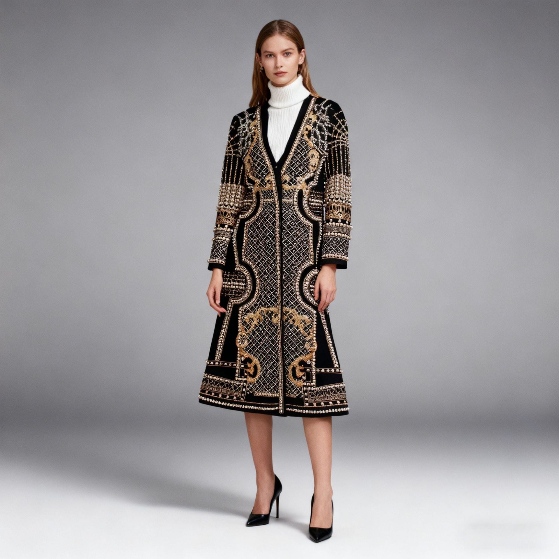 A model stands against a gray studio background wearing a long black embroidered coat with ornate gold and beige patterns, a white turtleneck, and black high heels.