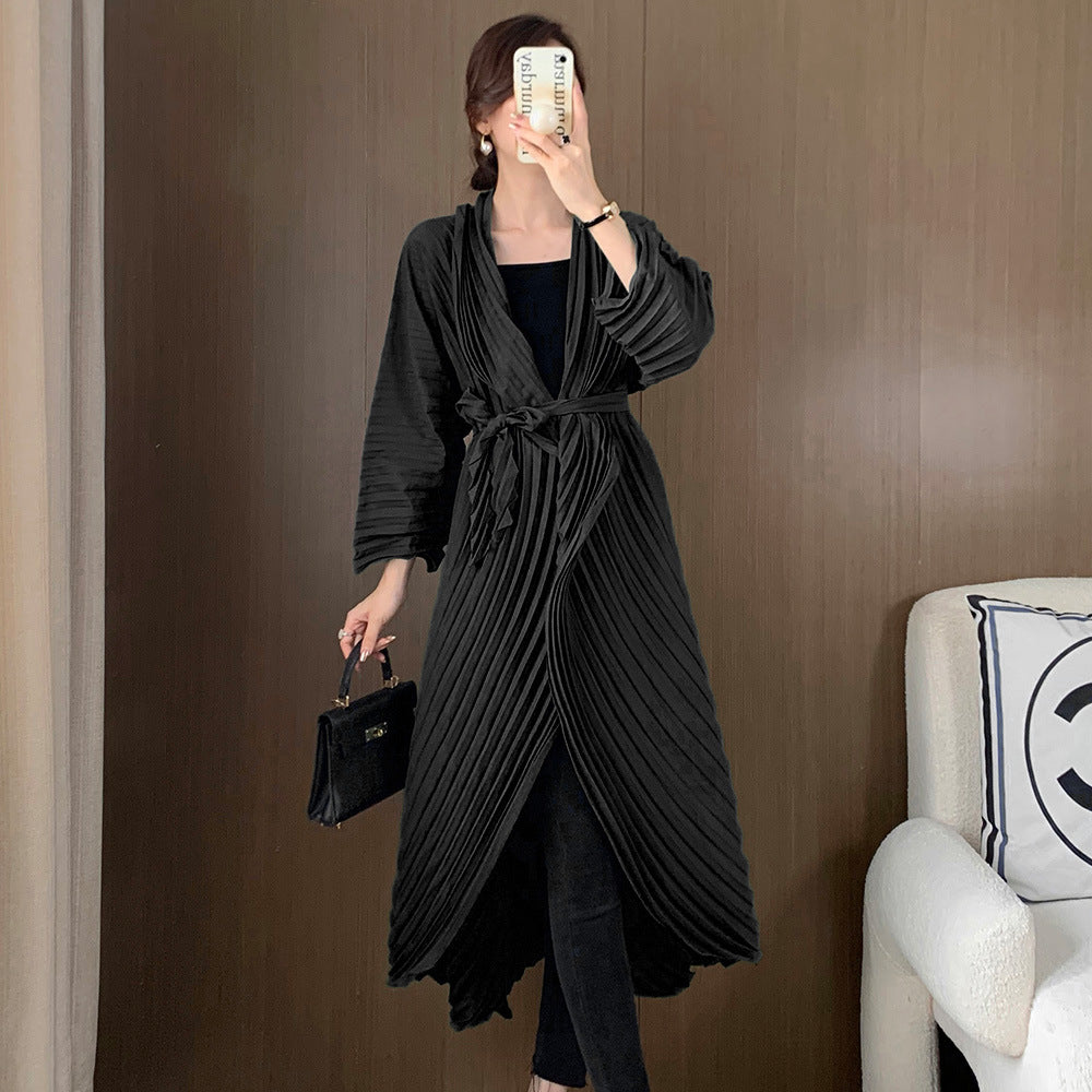 Woman wearing a long black coat holding a phone and a handbag against a brown wall.
