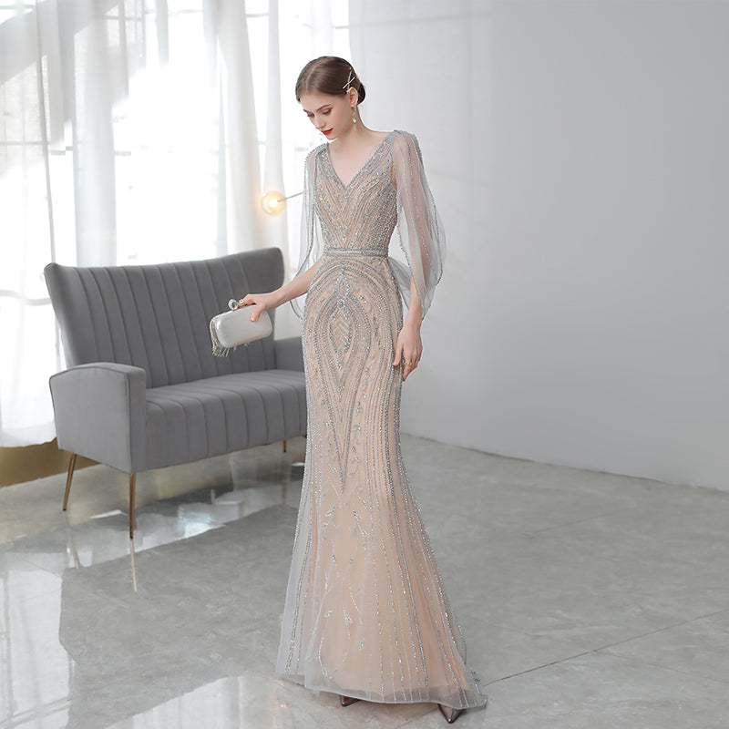 Woman in a long, elegant dress standing in a modern room with a gray sofa and white walls.